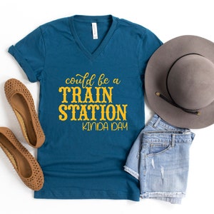 May include: A teal blue v-neck t-shirt with the text "could be a train station kinda day" in yellow lettering. The shirt is laid out on a white surface with a pair of brown sandals, a pair of denim shorts, and a gray fedora hat.