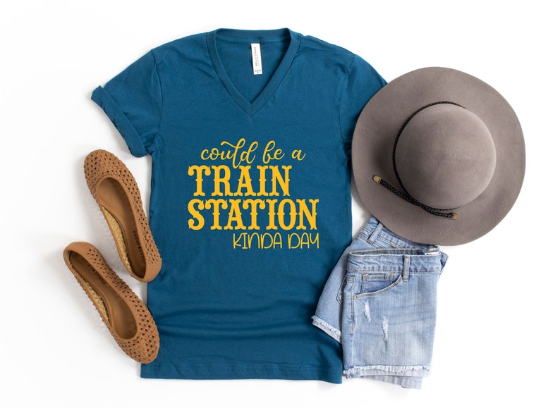 May include: A teal blue v-neck t-shirt with the text "could be a train station kinda day" in yellow lettering. The shirt is laid out on a white surface with a pair of brown sandals, a pair of denim shorts, and a gray fedora hat.