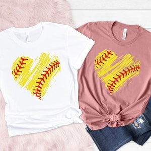 May include: Two t-shirts with a softball-shaped heart design. The heart is yellow with red stitching and a white background. One shirt is white and the other is pink.