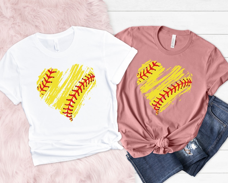 May include: Two t-shirts with a softball-shaped heart design. The heart is yellow with red stitching and a white background. One shirt is white and the other is pink.