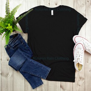May include: A black t-shirt with the text "Autumn Rain Clothing" printed on the front. The t-shirt is laid flat on a wooden surface with a pair of blue jeans and white trainers.