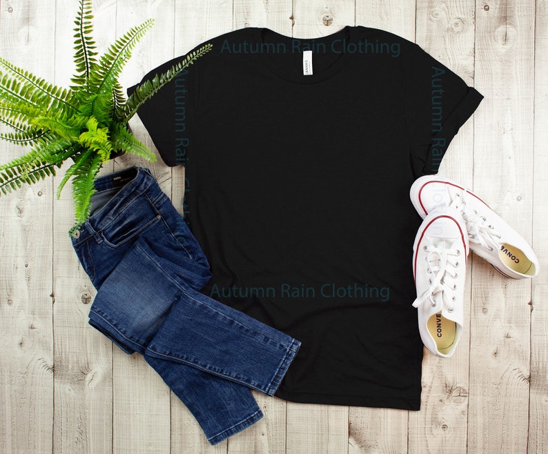 May include: A black t-shirt with the text "Autumn Rain Clothing" printed on the front. The t-shirt is laid flat on a wooden surface with a pair of blue jeans and white trainers.