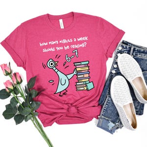 K&ouml;nnte beinhalten: Ein magentafarbenes T-Shirt mit dem Text "how many nights a week should you be reading?" und "6-7". Das Shirt zeigt eine Cartoon-Taube, die B&uuml;cher tr&auml;gt. Das Shirt wird mit Jeans und wei&szlig;en Slip-On-Schuhen kombiniert.