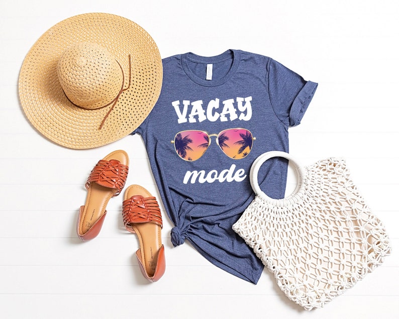 May include: A flat lay composition featuring a blue t-shirt with the words "VACAY mode" and sunglasses graphic. A straw hat, brown sandals, and a white woven tote bag are arranged around the shirt. The t-shirt is knotted at the side.