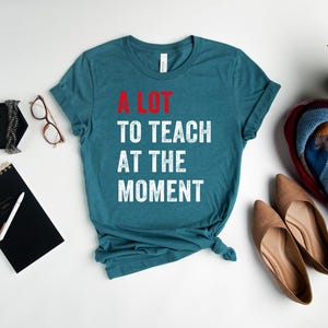 May include: A teal blue t-shirt with the text "A LOT TO TEACH AT THE MOMENT" in white with red accents. The text is in a distressed font.