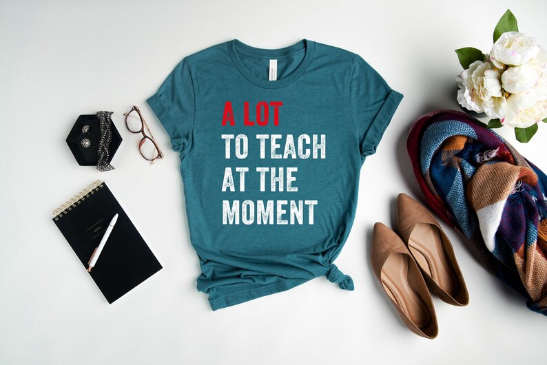 May include: A teal blue t-shirt with the text "A LOT TO TEACH AT THE MOMENT" in white with red accents. The text is in a distressed font.