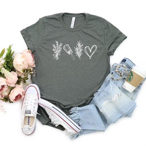 May include: A dark heather gray short-sleeve t-shirt featuring a white graphic print of a sprig of lavender, a salt shaker, a sprig of rosemary, and a heart outline. The shirt has a crew neckline and a standard fit with rolled sleeves.