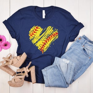 May include: A navy blue t-shirt with a yellow and red softball heart design. The shirt is laid flat on a white wooden surface with a pair of light brown heeled sandals and blue jeans.