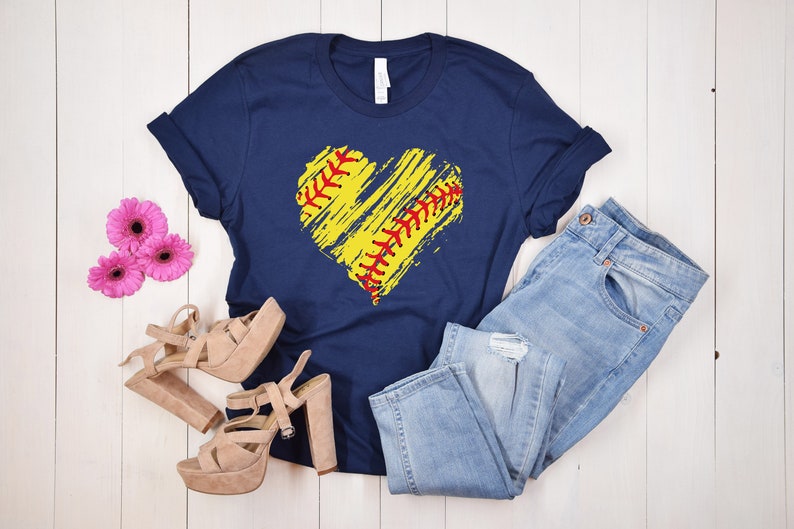 May include: A navy blue t-shirt with a yellow and red softball heart design. The shirt is laid flat on a white wooden surface with a pair of light brown heeled sandals and blue jeans.