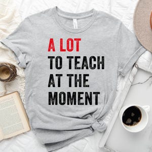 May include: A gray t-shirt with the text "A LOT TO TEACH AT THE MOMENT" in black with the word "LOT" in red.
