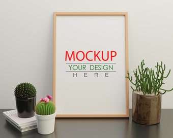 以下が含まれることがあります： 白い背景に「MOCKUP YOUR DESIGN HERE」というテキストが書かれた木製フレームのモックアップ。フレームは黒い表面に置かれ、サボテン2つと多肉植物1つを含む3つの鉢植えがあります。