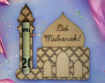 May include: Wooden cutout of a mosque with a slot for cash, decorated with a geometric pattern. The mosque has a dome with the words "Eid Mubarak!" written on it. The cutout is on a light blue background with pink and gold confetti.