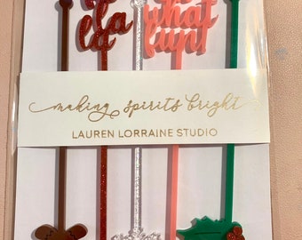 May include: Set of six festive cocktail stirrers with various holiday-themed designs. The stirrers feature a gingerbread man, snowflake, holly, and the phrases "la la la" and "oh what fun". The packaging includes the text "making spirits bright" and "Lauren Lorraine Studio".