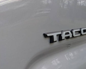 May include: A close-up of a gray pickup truck with the word "TACOMA" in black lettering on the side.