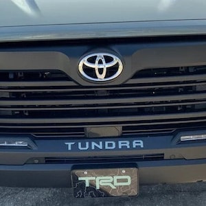 May include: A gray Toyota Tundra pickup truck with a black grille and a chrome Toyota logo. The truck has a black "Tundra" lettering on the front bumper and a black "TRD" license plate.
