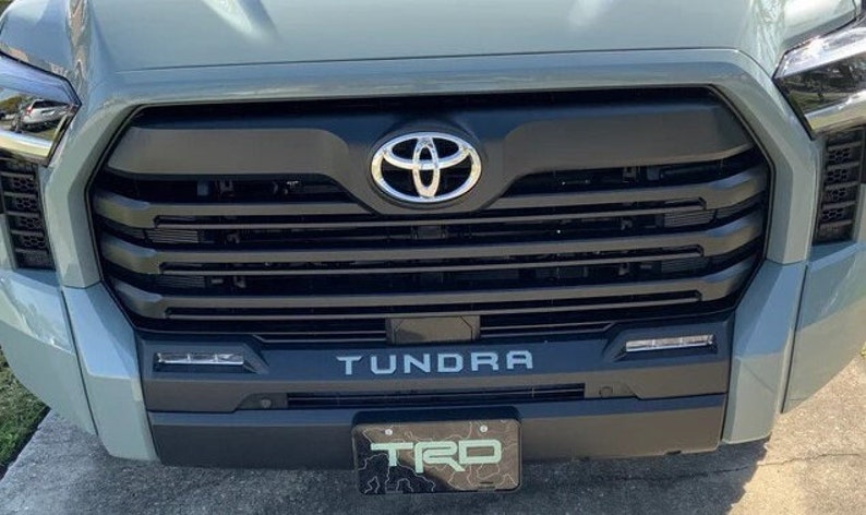 May include: A gray Toyota Tundra pickup truck with a black grille and a chrome Toyota logo. The truck has a black "Tundra" lettering on the front bumper and a black "TRD" license plate.