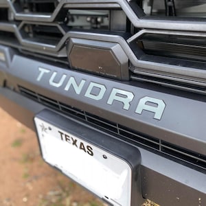May include: Close-up of a black truck grille with the word "Tundra" in gray lettering. A black license plate with the word "Texas" is visible below the grille.