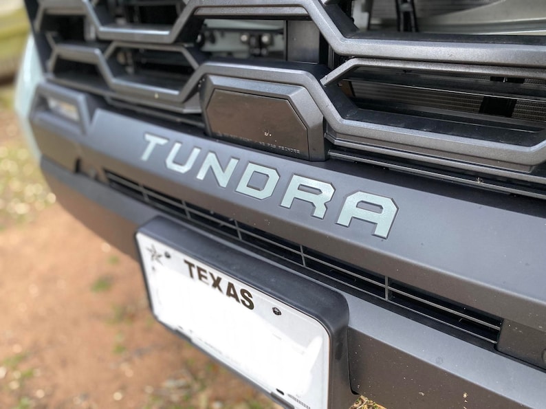 May include: Close-up of a black truck grille with the word "Tundra" in gray lettering. A black license plate with the word "Texas" is visible below the grille.