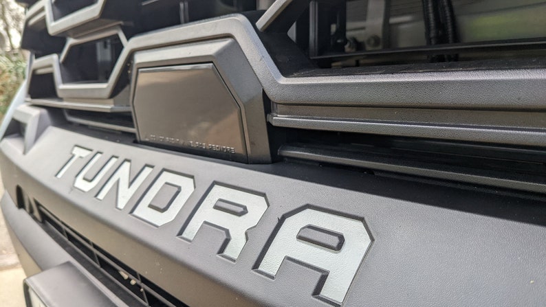 May include: Close-up of a black truck grille with the word "TUNDRA" in silver lettering.