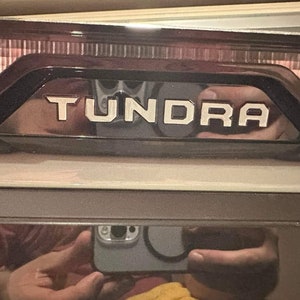 May include: A close-up of a black truck tailgate with the word "Tundra" in white lettering.