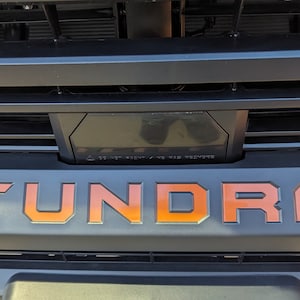 May include: A close-up of a black truck grille with the word "TUNDRA" in orange lettering.