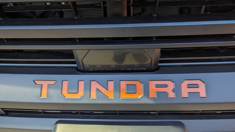 May include: A close-up of a black truck grille with the word "TUNDRA" in orange lettering.