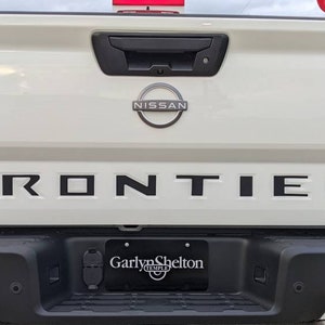 May include: White Nissan Frontier SV pickup truck with black lettering on the tailgate that reads "FRONTIER". The truck has a black bumper with a license plate that reads "Garlyn Shelton".