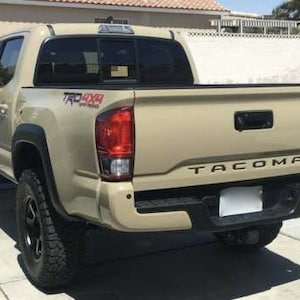 May include: A beige Toyota Tacoma pickup truck with black trim and a black rear bumper. The truck has a black "TACOMA" emblem on the tailgate and a "TRD 4X4" emblem on the side. The truck is parked on a paved surface.