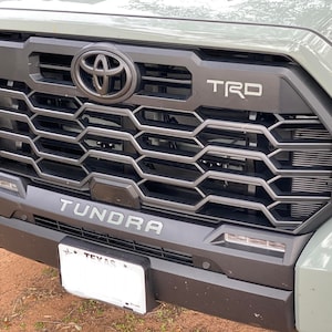 Puede incluir: Primer plano de una camioneta Toyota Tundra gris con una parrilla negra y un emblema "TRD" negro. La camioneta tiene un emblema "Tundra" negro debajo de la parrilla y una placa de matrícula de Texas.