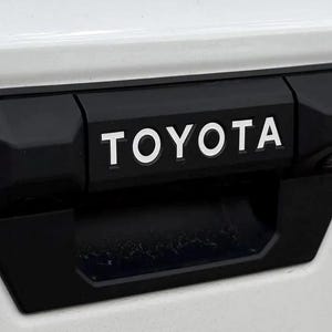 May include: A black plastic truck tailgate handle with a white "TOYOTA" logo.