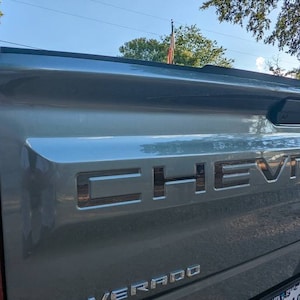 Peut inclure: L'arrière d'un pick-up Chevrolet Silverado gris avec le logo Chevrolet sur le hayon. Le pick-up a un pare-chocs noir et une poignée de hayon noire.