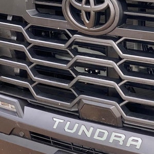 May include: Close-up of a black Toyota Tundra grille with a chrome Toyota logo and the word "Tundra" in silver lettering.