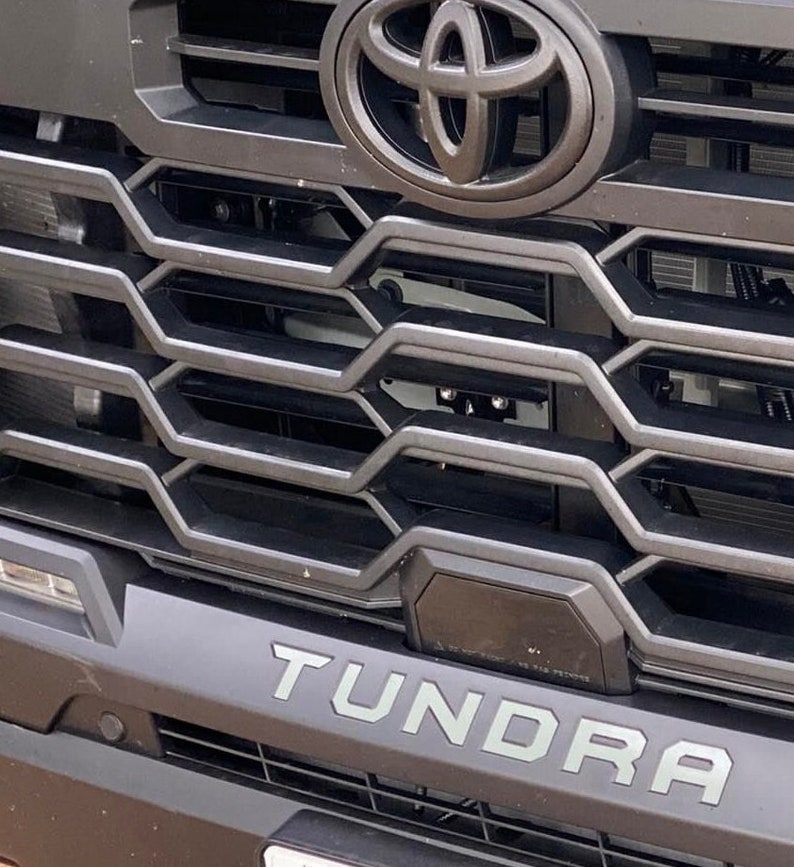 May include: Close-up of a black Toyota Tundra grille with a chrome Toyota logo and the word "Tundra" in silver lettering.