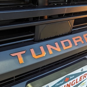 May include: A close-up of a black truck grille with a large "TUNDRA" emblem in orange and red lettering. The truck is parked in a parking lot.
