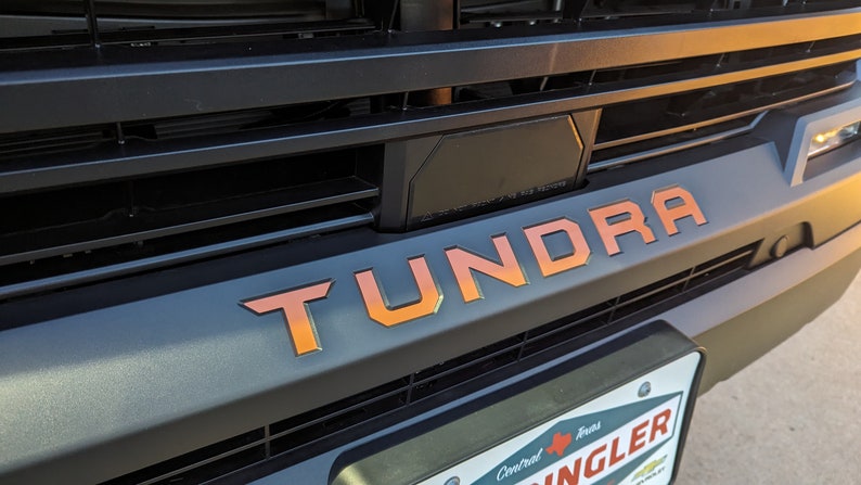 May include: A close-up of a black truck grille with a large "TUNDRA" emblem in orange and red lettering. The truck is parked in a parking lot.