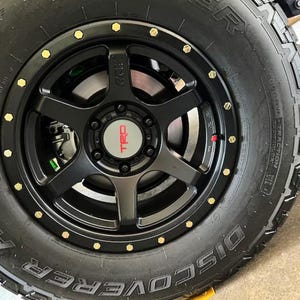 Könnte beinhalten: Schwarzer Reifen mit goldenen Radmuttern und einem schwarzen Rad mit einem roten "TRD"-Logo. Der Reifen hat die Aufschrift "DISCOVERER" auf der Seite.