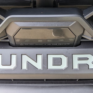 May include: A close-up of a black truck grille with the word "TUNDRA" in silver lettering. The grille has a black plastic trim piece with a warning label that reads "DO NOT PAINT / NO PAS DE PEINTURE".
