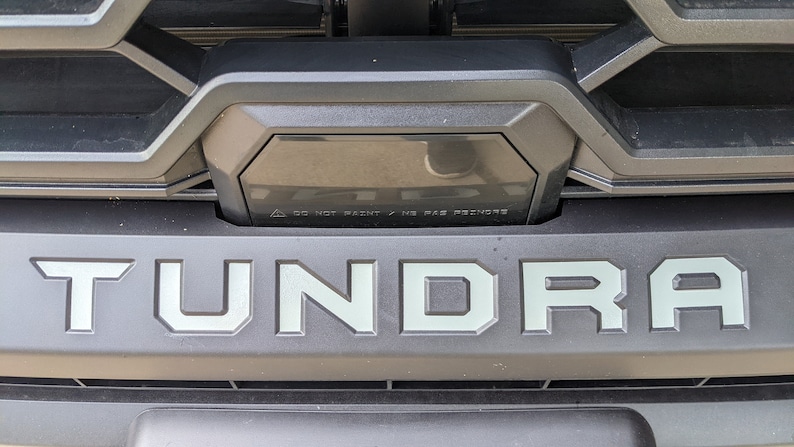 May include: A close-up of a black truck grille with the word "TUNDRA" in silver lettering. The grille has a black plastic trim piece with a warning label that reads "DO NOT PAINT / NO PAS DE PEINTURE".