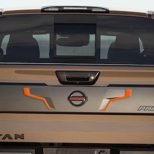 May include: Rear view of a brown Nissan Titan pickup truck with a black tailgate and the words "TITAN" and "PRO-4X" in black lettering. The truck has orange accents on the tailgate.
