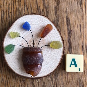 Op de afbeelding: Een handgemaakte houten schijf met een bloemstuk van zeeglas. De bloem heeft een bruine zeeglasvaas en kleurrijke zeeglasblaadjes in groen, geel, blauw en oranje. Een Scrabble-tegel met de letter "A" staat ernaast.