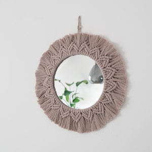 May include: A circular mirror with a beige macrame frame and a fringed edge. The mirror is suspended on a white wall. The reflection shows a green plant and a grey pendant light.