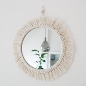Può includere: Uno specchio rotondo con una cornice in macramé bianca e frange. Lo specchio è appeso a una parete bianca e riflette un lampadario e una pianta.