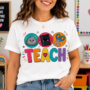 May include: A white short-sleeve t-shirt featuring a colourful graphic with the word TEACH in large letters, where the A is replaced by a pink heart. Above the text are three circular icons depicting a grey rock, a black notebook, and blue scissors.