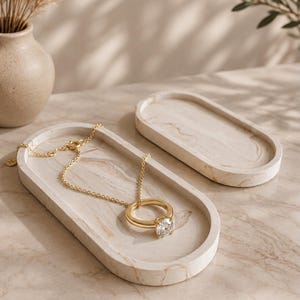 Può includere: Due vassoi di forma ovale con un motivo marmorizzato beige e bianco. Un vassoio contiene una collana a catena color oro con un ciondolo circolare caratterizzato da una pietra trasparente sfaccettata. I vassoi hanno bordi lisci e rialzati.
