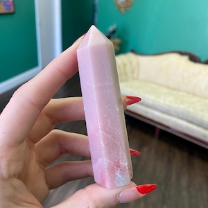 May include: A pink and white crystal point with a faceted shape. The crystal is about 4 inches long and has a smooth, polished surface.