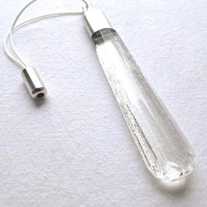 May include: A clear, elongated glass pendant with a textured surface and a silver-coloured metal cap. A thin, white cord is attached to the pendant, leading to a small, silver-coloured metal bead. The pendant is displayed on a white background.