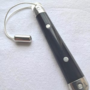 May include: A black pocket knife with silver detailing and a lanyard. The knife features a dark handle with two silver rivets and silver end caps. The lanyard is a thin, white cord with a silver bead.