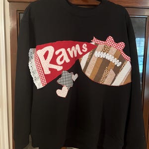 May include: Black sweatshirt with a decorative design featuring the word "Rams" in white on a red banner. The design includes a football shape with various patterned fabrics and heart shapes.