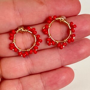 May include: A pair of gold-toned hoop earrings adorned with vibrant red beads. The earrings have a circular design, with the red beads evenly spaced around the hoops. The earrings are displayed against a neutral background.