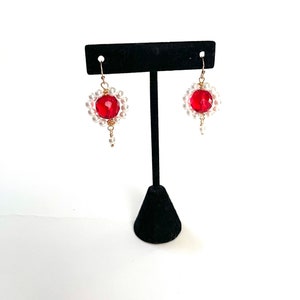 May include: A pair of dangle earrings featuring a large, faceted red bead surrounded by small white pearls. The earrings have gold-colored wire accents and a small pearl drop. The jewelry is displayed on a black stand against a white background.